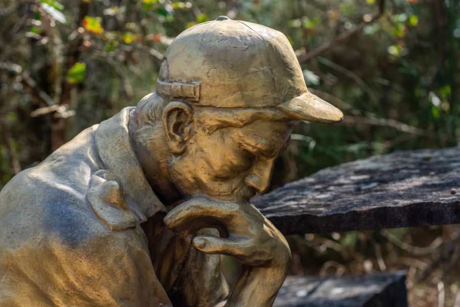 Statua dorata di un uomo con cappellino, pensieroso, con mano sul mento, seduto all'aperto.