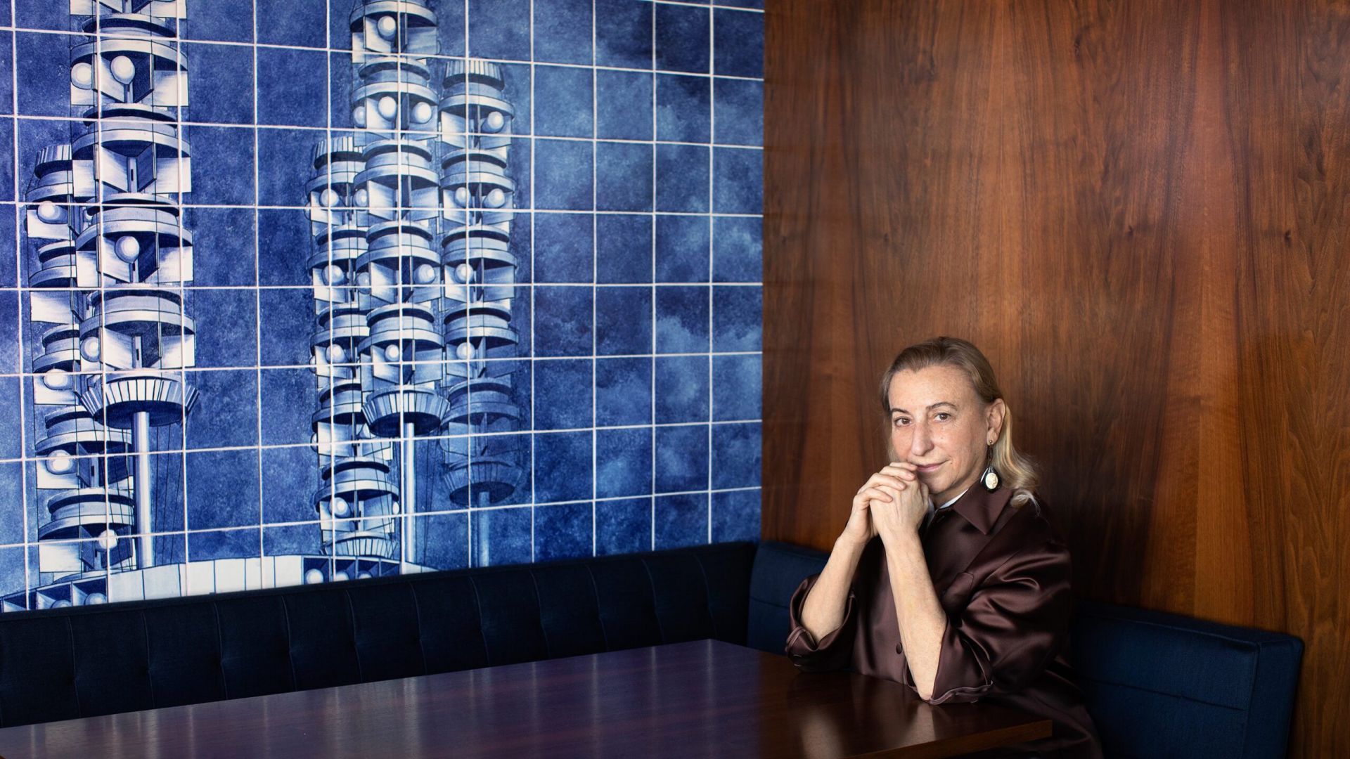 La collezionista Miuccia Prada, seduta su una panca blu, davanti a un muro di piastrelle blu che compongono un'opera d'arte e parete di legno scuro. La signora Prada compare nella Top 200 collezionisti ARTnews anche per il 2026