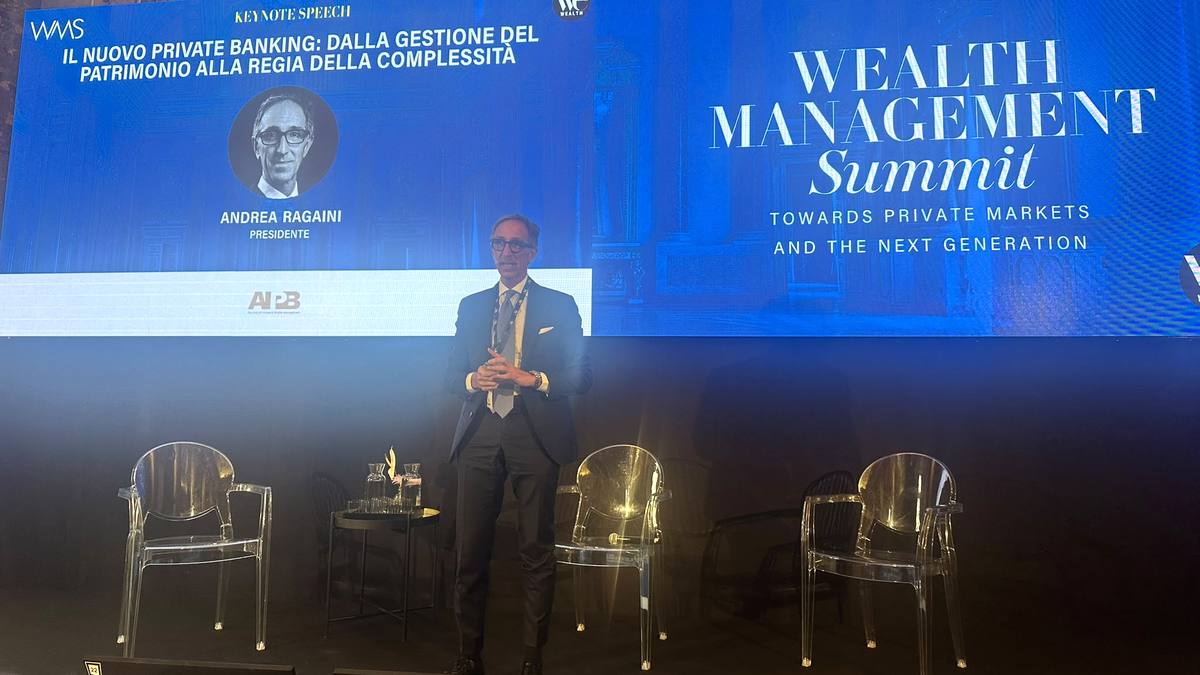 Uomo in abito parla sul palco, con sfondo blu e logo "Wealth Management Summit".