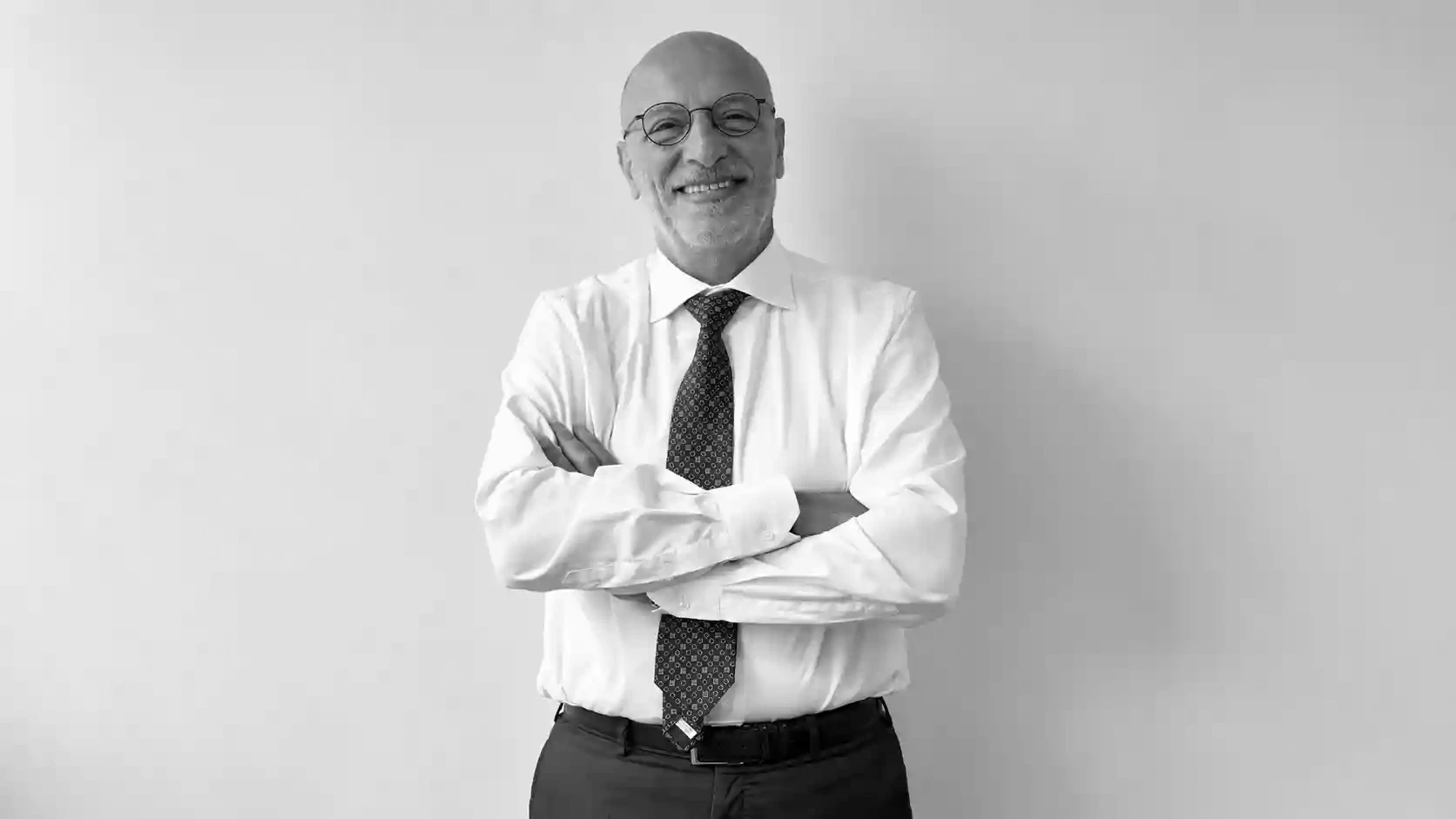 Uomo sorridente con occhiali, camicia bianca e cravatta, braccia conserte.