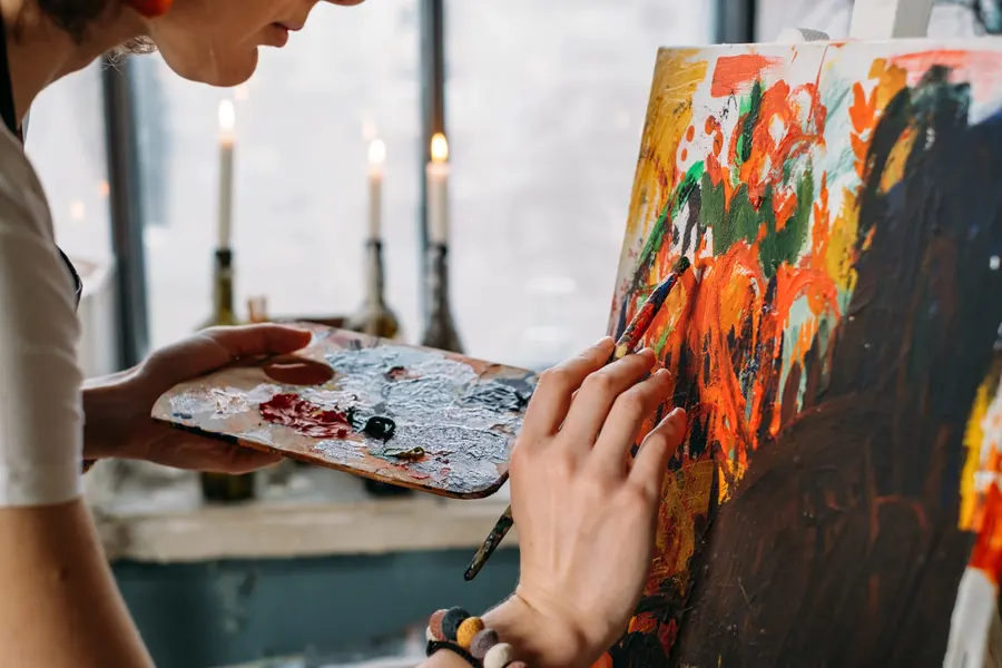 Artista dipinge su tela con tavolozza e pennello, candele sullo sfondo.