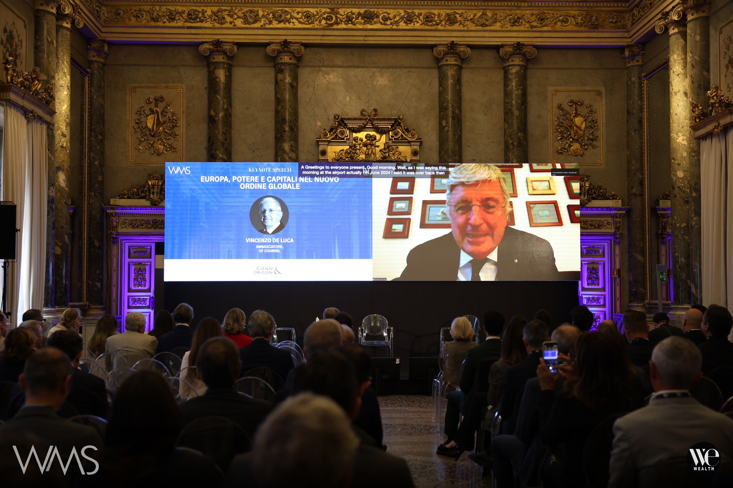 Vincenzo De Luca con occhiali parla in videochiamata, pubblico in sala ascolta durante il terzo Wealth management Summit di We Wealth.
