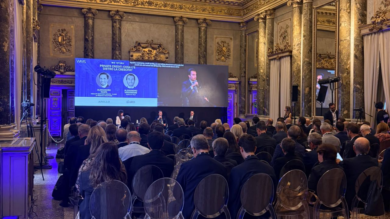 Presentazione su schermo gigante con relatori e pubblico in sala.