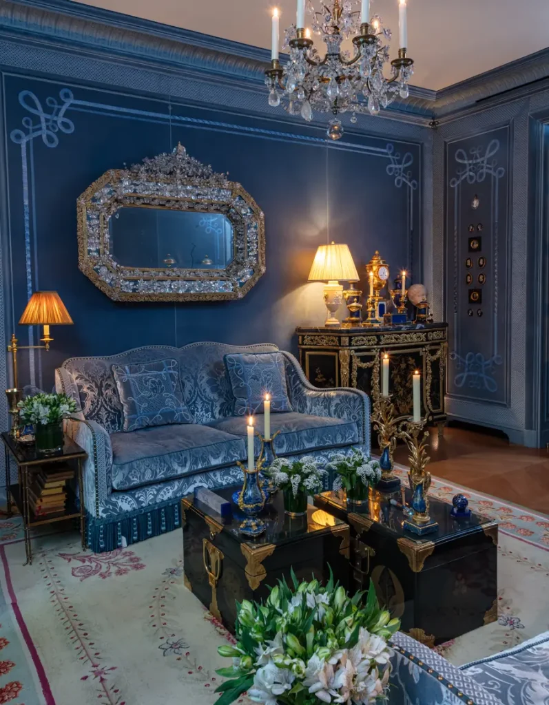 Salotto blu con divano, specchio ornato, candelabri dorati e due bauli neri.