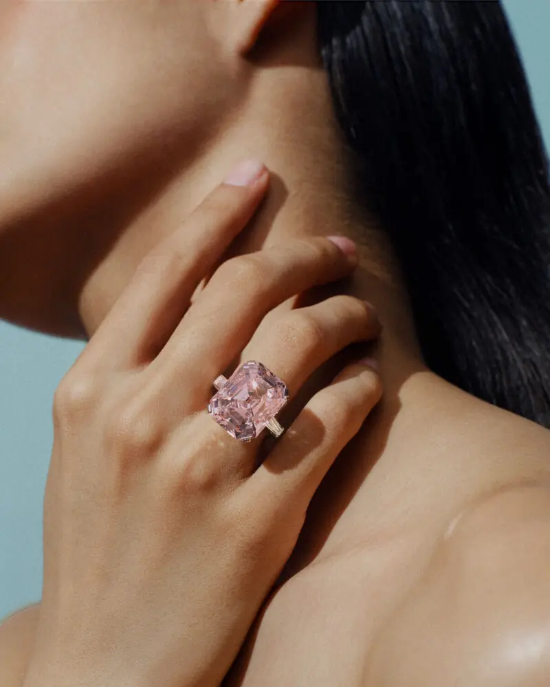 Mano con anello con grande pietra rosa sfaccettata, dita che sfiorano il collo.