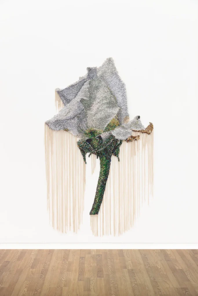 Fiore stilizzato grigio e verde con frange pendenti su muro bianco e pavimento in legno.