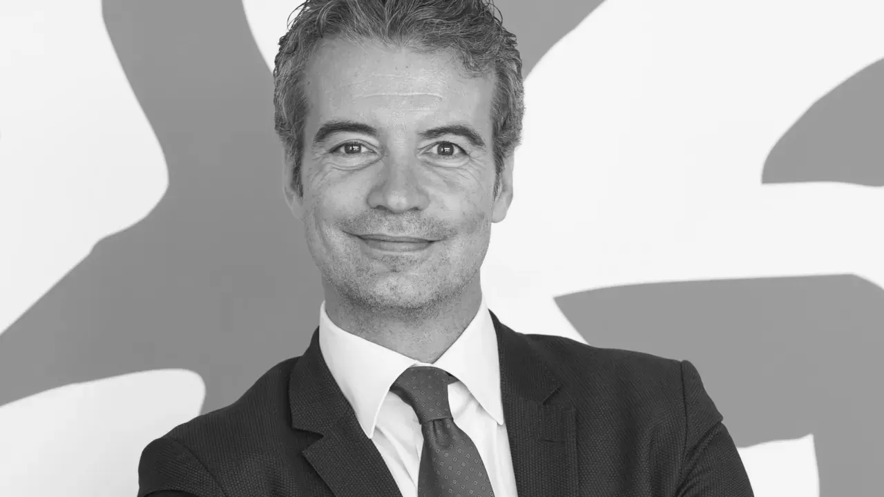 Uomo sorridente in abito scuro e cravatta, con sfondo astratto in bianco e nero.