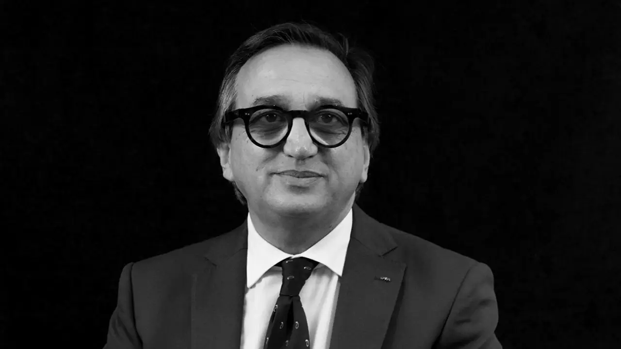 Uomo con occhiali, abito scuro e cravatta, su sfondo nero.
