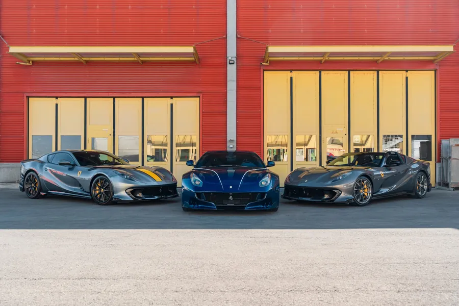 Tre Ferrari sportive, una blu al centro, due grigie ai lati, davanti a porte gialle.