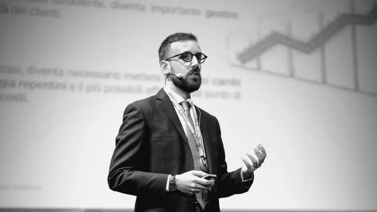 Uomo con occhiali e barba parla, indossa giacca e cravatta, con sfondo di grafici.