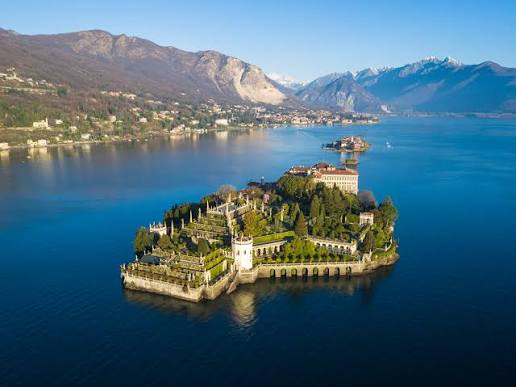 Isola con palazzo e giardini terrazzati su lago blu, montagne sullo sfondo.