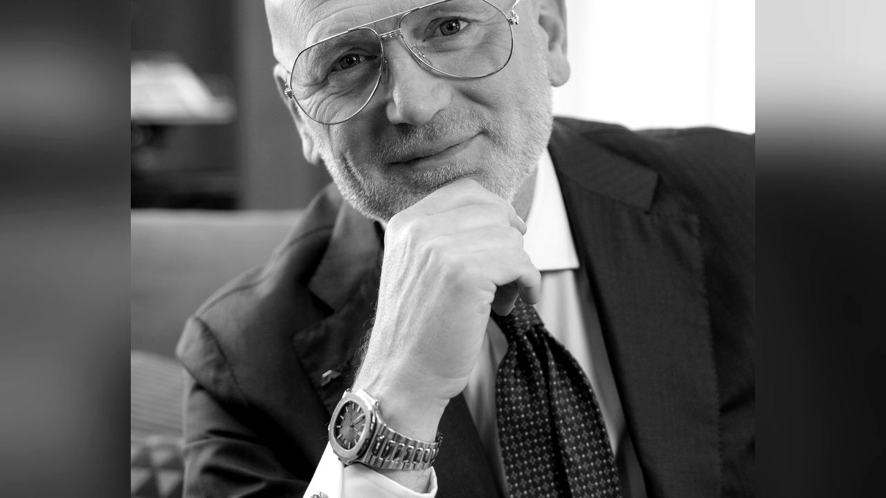Uomo con occhiali, barba, giacca e cravatta, con mano sul mento.