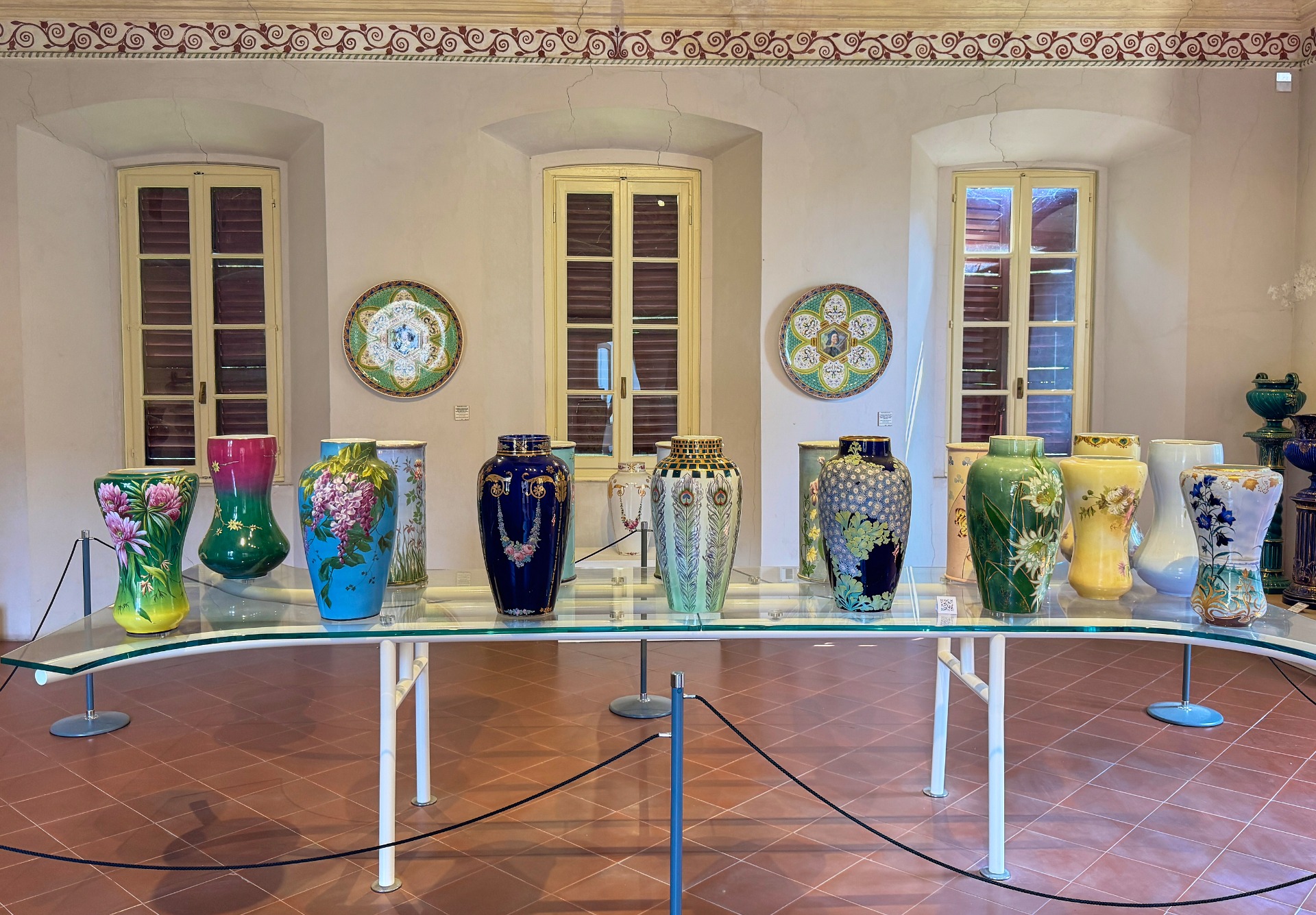 Vasi decorati in ceramica su un tavolo di vetro, con finestre e decorazioni murali sullo sfondo.