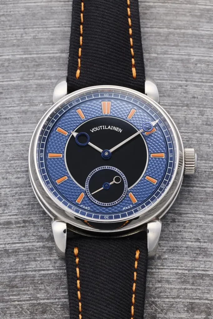 Orologio da polso con quadrante blu guilloché, lancette arancioni e cinturino nero con cuciture arancioni.