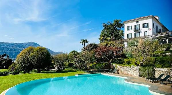 Villa bianca con piscina, alberi verdi e montagne sullo sfondo.