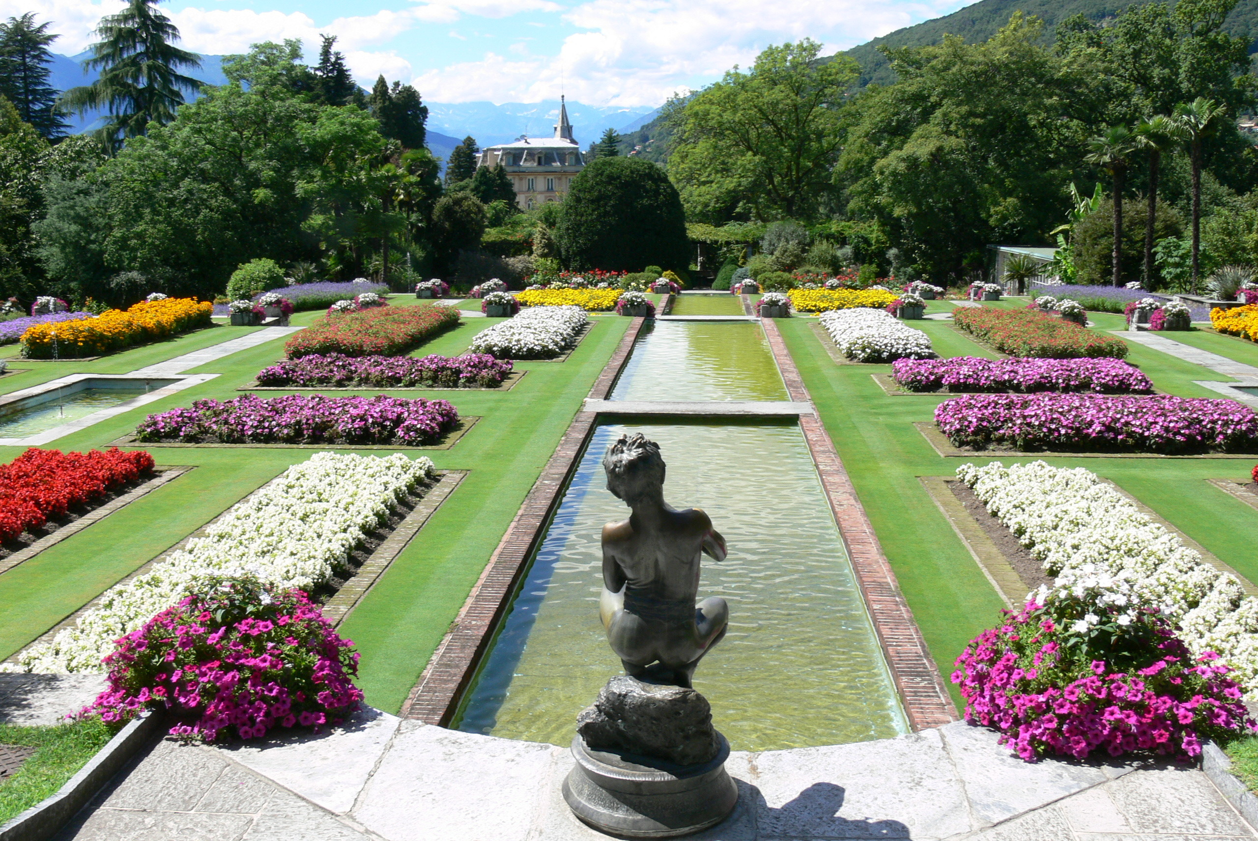 Giardino con statue, fontane, aiuole fiorite colorate e montagne sullo sfondo.