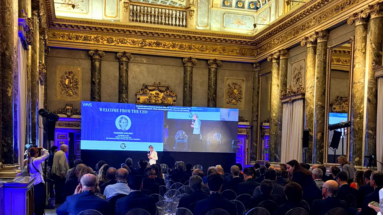 Presentazione su grande schermo in una sala sontuosa con pubblico seduto.